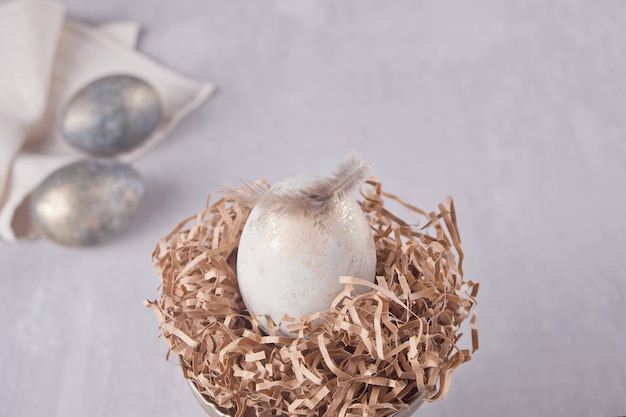 Weißes Ei Ostern in einem Nest und zwei silbernen Eiern auf einem grauen Hintergrund mit Federn