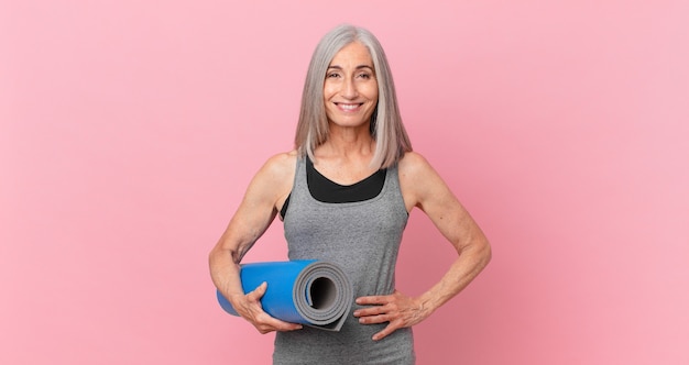 Weißes Haar Frau mittleren Alters glücklich lächelnd mit einer Hand auf der Hüfte und selbstbewusst und hält eine Yogamatte. Fitnesskonzept