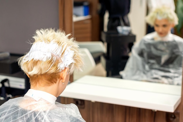 Weißes Haar mit Haarfärbemittel der jungen kaukasischen blonden Frau färben, die in einem Friseursalon sitzt