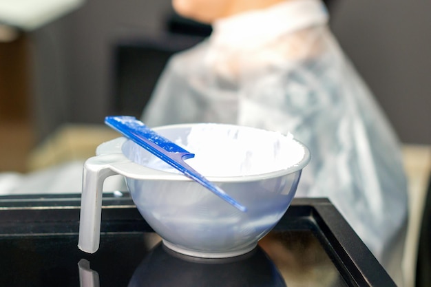 Weißes Haarfärbemittel im Behälter auf dem speziellen Tisch auf weiblichem Kundenhintergrund im Friseursalon