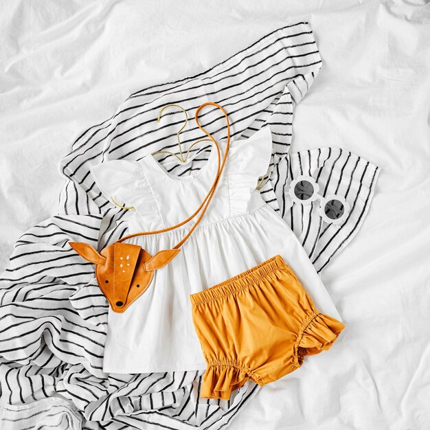 Weißes Kleid, orangefarbene Shorts mit Kinderhandtasche und Sonnenbrille. Set Babykleidung und Accessoires für den Sommerurlaub auf dem Bett. Mode Kinder Outfit. Flache Lage, Ansicht von oben