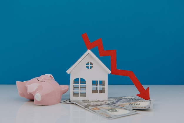 Weißes Modell des Hauses und kaputtes Sparschwein mit Pfeil nach unten Fallende Immobilienmarktpreise