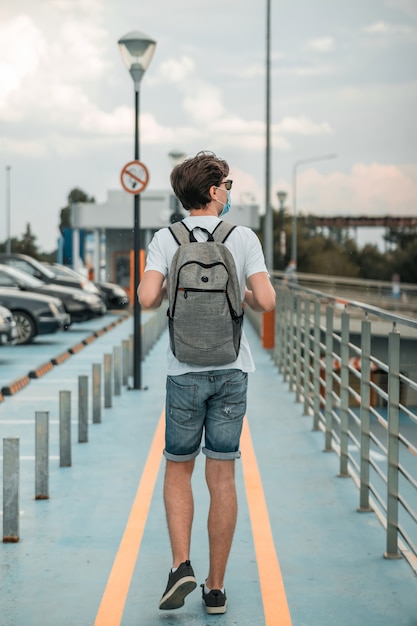 Weißes T-Shirt des jungen Erwachsenen i, Sonnenbrille, Jeansshorts, schwarzes Schuhwerk, OP-Maske und Rucksack, die auf dem Parkplatz gehen