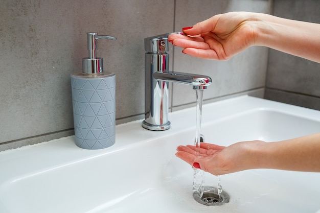 Weißes Waschbecken mit silbernem Wasserhahn im Badezimmer. Graue Dose mit Flüssigseife für die Hände. Aufdrehen des Leitungswassers, persönliche Händehygiene. Händewaschen unter fließendem Wasser