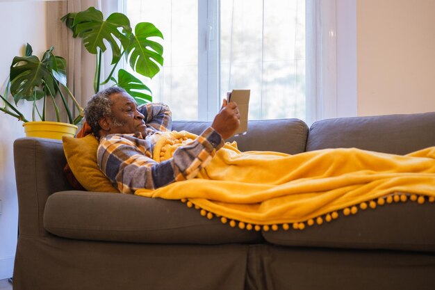Weißhaariger älterer Mann, der auf dem Sofa liegt und Tablet liest und sich zu Hause ausruht