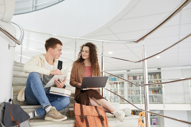 Weitwinkelansicht bei zwei Studenten, die auf Treppen in der Universitätsbibliothek sitzen und Laptop während der Arbeit an Hausaufgaben verwenden,