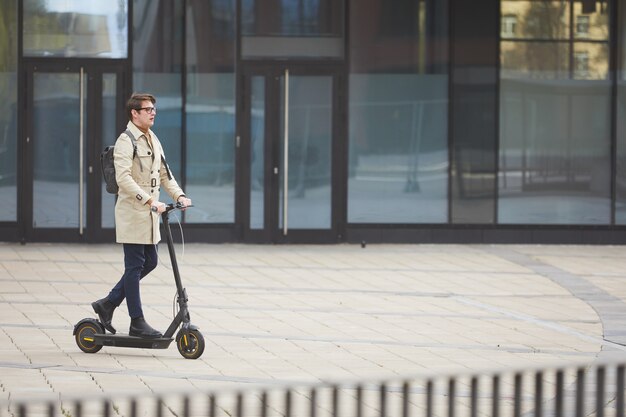 Weitwinkelporträt des modernen jungen Geschäftsmannes, der Elektroroller reitet, während zur Arbeit mit städtischen Stadtgebäuden im Hintergrund, Kopierraum pendelt