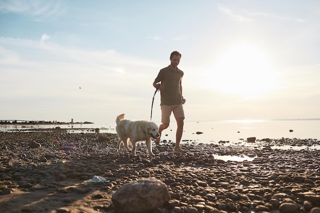 Weitwinkelporträt in voller Länge eines glücklichen erwachsenen Mannes, der mit Hund am Wasser bei Sonnenuntergang Kopienraum läuft