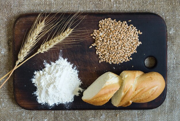 Weizenähren, Körner, Mehl und geschnittenes Brot auf einem Küchenbrett auf einem Entlassungshintergrund. Ansicht von oben