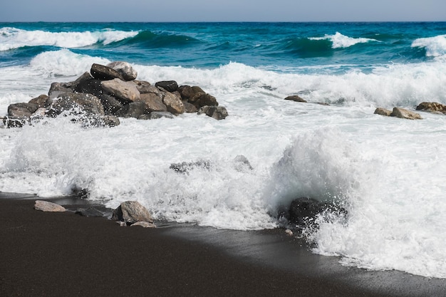 Wellen am Strand an windigen Tagen. Strand von Perissa mit schwarzem Vulkansand auf der Insel Santorini, Griechenland. Blaues Meer und blauer Himmel