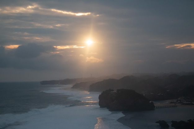 Wellen, die gegen Felsen und Sandstrand bei Sonnenuntergang schlagen, mit Sonnenstrahlen und Wolken am Himmel