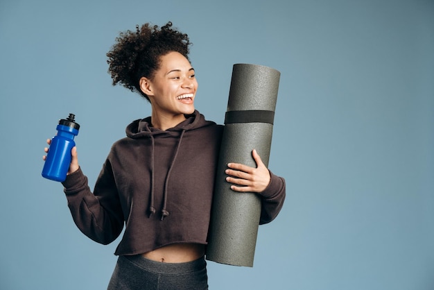 Wellness. Lächelnde tausendjährige Sportlerin, die Yogamatte und Flasche mit Wasser hält, während sie isoliert auf blauem Studiohintergrund steht. Positive junge Frau bereit für ihr morgendliches Training