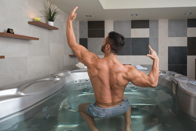 Wellness Spa hombre flexionando los músculos en la bañera de hidromasaje Jacuzzi interior en el lujoso Resort Spa Retreat Guapo joven modelo masculino de pie fuerte en el agua cerca de la piscina en vacaciones de viaje