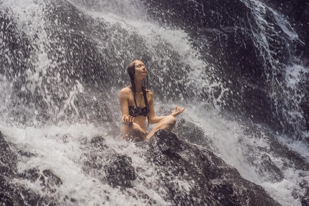 Wellness-Spa-Urlaub und Yoga-Meditationskonzept Junge Frau sitzt in Lotusposition auf dem Felsen im tropischen Wasserfall