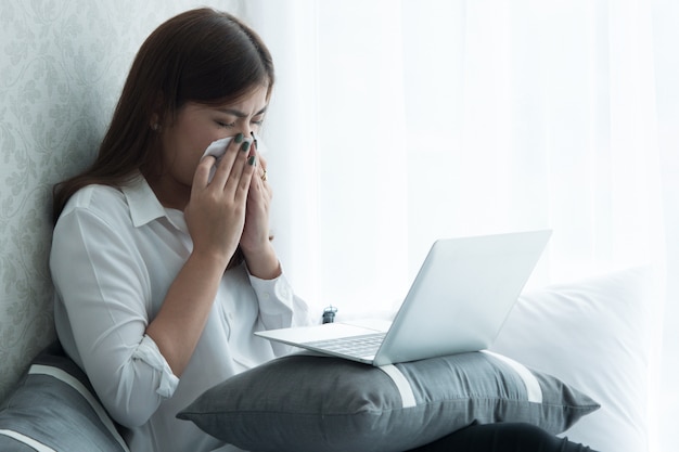 Wenn eine Frau Fieber hat, niest sie, während sie einen Laptop im Schlafzimmer benutzt.