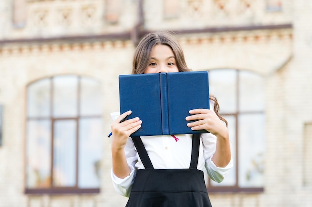 Wer hat dieses Buch ausgewählt? Entzückendes kleines Kind, das Gesicht mit Buch bedeckt. Nettes Lesebuch des kleinen Mädchens im Freien. Sie sind nur ein Buch von guter Laune entfernt.