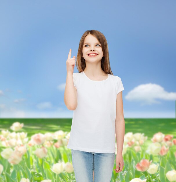 Werbe-, Kindheits-, Natur-, Gesten- und Personenkonzept - lächelndes Mädchen im weißen T-Shirt, das mit dem Finger nach oben über den Hintergrund der Blumenwiese zeigt