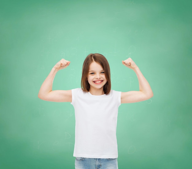 Werbung, Geste, Schule, Bildung und Menschen - lächelndes kleines Mädchen in weißem, leerem T-Shirt mit erhobenen Armen über grünem Bretthintergrund