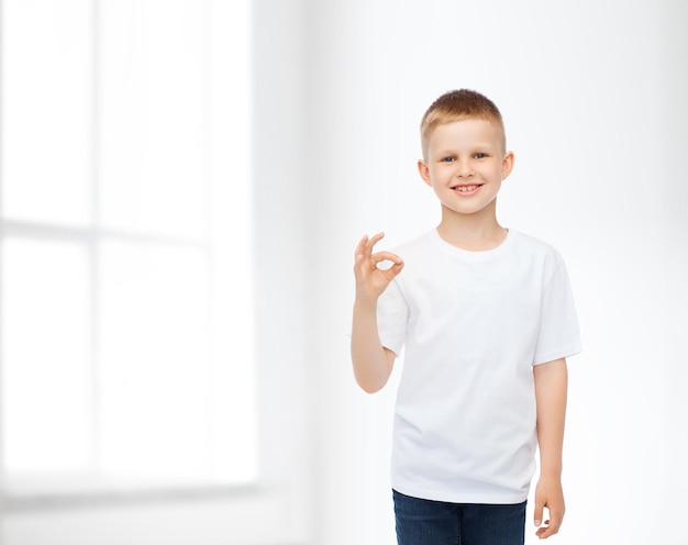 werbung, leute, zuhause und kindheitskonzept - lächelnder kleiner junge im weißen leeren t-shirt, das ok geste über weißem raumhintergrund macht