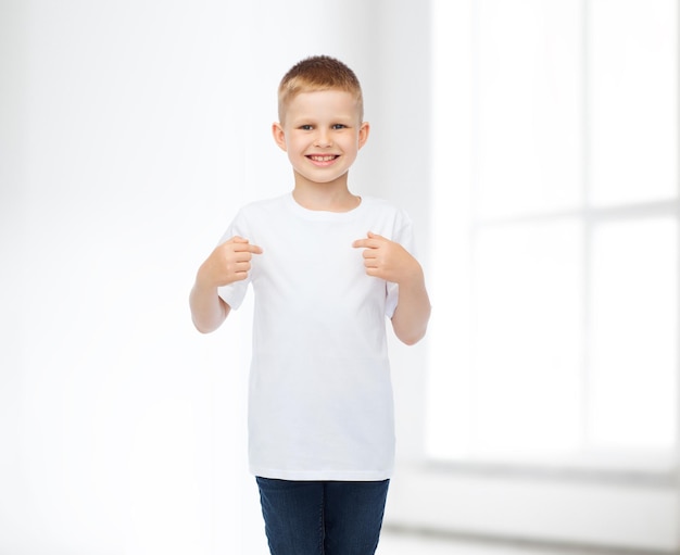 Werbung, Menschen, Gesten und Kindheitskonzept - lächelnder Junge im weißen, leeren T-Shirt, der mit dem Finger auf den Hintergrund des weißen Raums zeigt