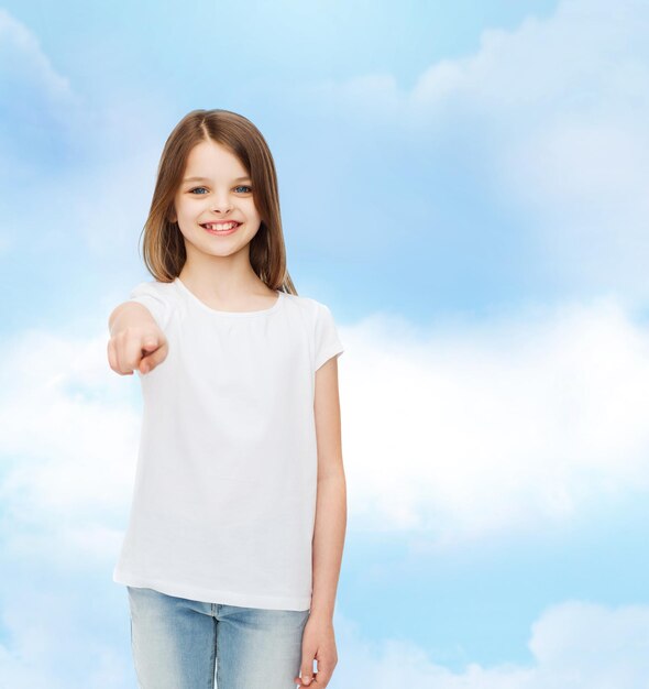 Werbung, Traum, Kindheit, Gesten und Menschen - lächelndes kleines Mädchen im weißen T-Shirt, das mit dem Finger auf dich über bewölktem Himmelshintergrund zeigt