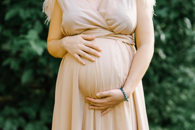 Werdende Mutter im beige schönen Kleid mit Federn hält ihre Hände auf ihrem schwangeren Bauch