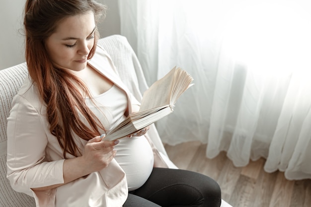 Werdende Mutter in den letzten Monaten der Schwangerschaft mit einem Buch auf der Couch zu Hause am Fenster.