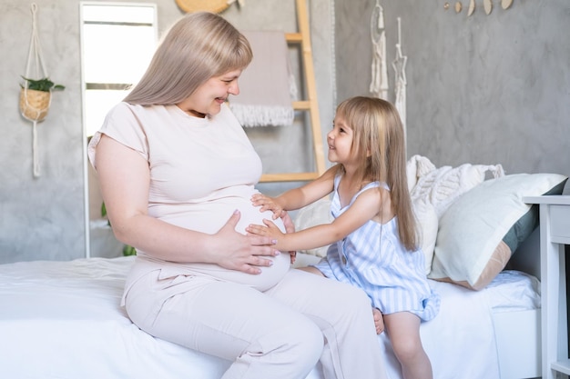 Werdende Mutter sitzt auf dem Bett mit kleiner Tochter, die den Bauch streichelt