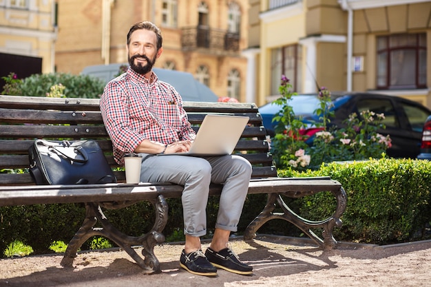 Werktags. Fröhlicher gutaussehender Mann, der auf der Bank sitzt, während er seinen Laptop benutzt