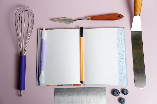 Werkzeuge zum Erstellen und Dekorieren eines Kuchens, verschiedene Spachtel und Sahnedüsen mit einem offenen Notizbuch und einem Stift für Text und Rezepte auf violettem Hintergrund
