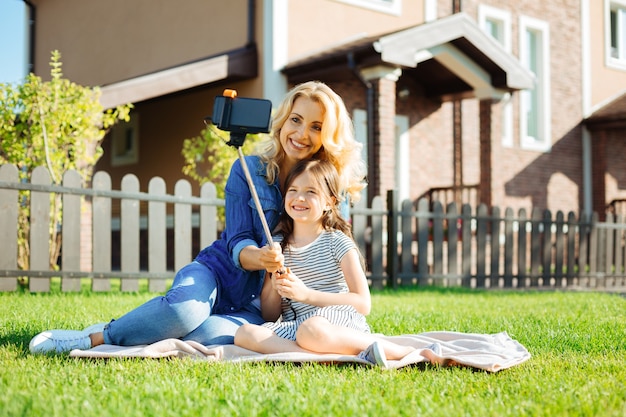 Wertvolle Momente. Entzückendes kleines Mädchen, das neben ihrer angenehmen Mutter auf dem Teppich sitzt und zusammen mit einem Selfie-Stick ein Selfie mitnimmt