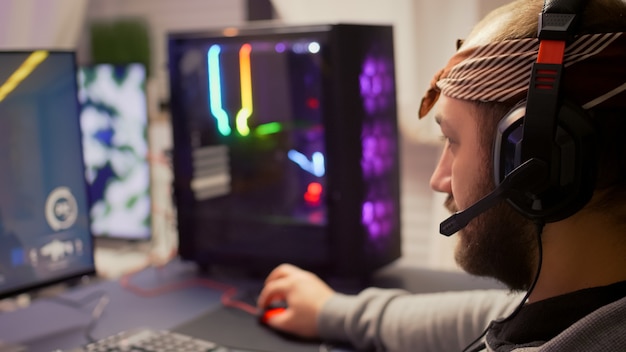 Wettbewerbsfähiges Cyber-Pro-Gamer-Streaming mit Mikrofon, das Ego-Shooter-Videospiele während des virtuellen Turniers spielt. Spieler mit professionellem RGB-System-Desktop im Gaming-Heimstudio spät am Abend