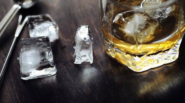 Whisky con hielo con vaso. Cubos de hielo sobre una mesa de madera y un vaso con alcohol fuerte refrigerado. Mesa con whisky americano, bourbon y cartas.