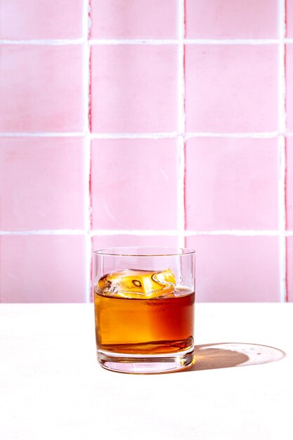 Whisky o bourbon en un vaso de rocas con un gran cubo de hielo con luz dura y sombras duras espacio de copia de fondo de azulejo rosa