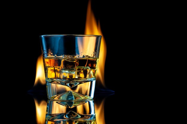 Whisky oder Bourbon in einem transparenten Glas mit Eiswürfeln auf einem schwarzen mit Feuer