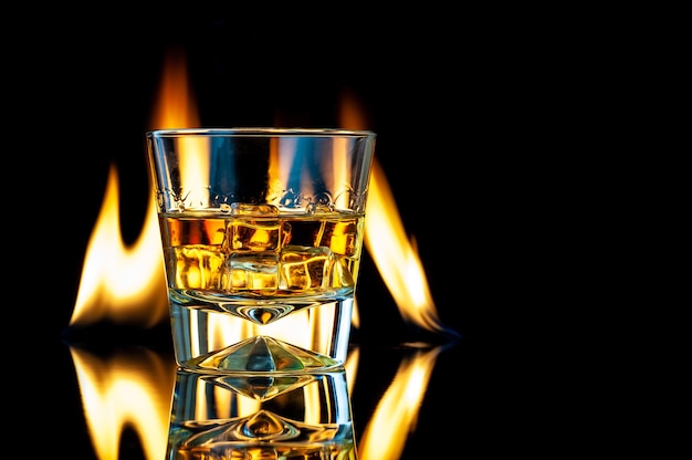 Whisky oder Bourbon in einem transparenten Glas mit Eiswürfeln auf einem schwarzen mit Feuer