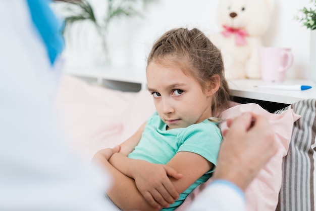 Widerstrebendes krankes kleines Mädchen, das Löffel mit Medizin betrachtet, die vom Arzt gehalten wird, während im Bett zu Hause sitzen