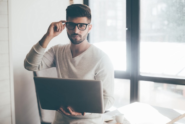 Wie kann ich dir helfen? Selbstbewusster junger Mann, der Laptop hält und seine Brille justiert, während er sich im Büro am Schreibtisch lehnt und in die Kamera schaut