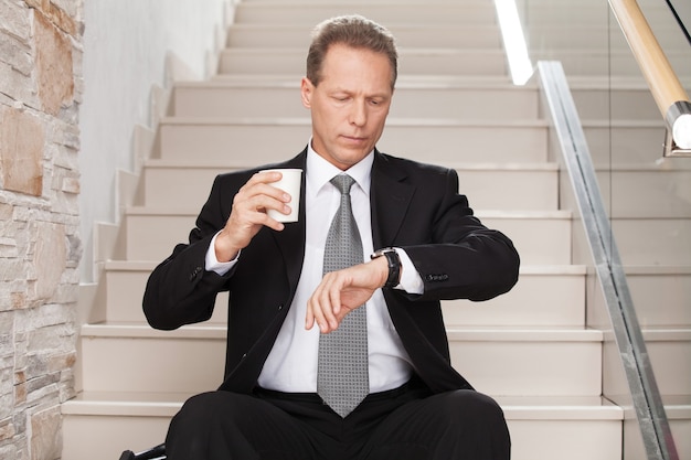Wieder spät. besorgter reifer mann in formeller kleidung, der eine kaffeetasse hält und eine zeit überprüft, während er auf der treppe sitzt