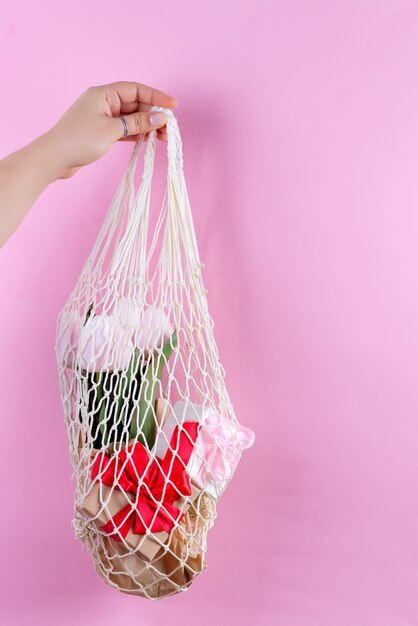 Wiederverwendbare Einkaufstaschen mit Geschenkboxen aus Papier und frischen Frühlingstulpen blühen in der Hand der Frau vor einem rosa Hintergrund.