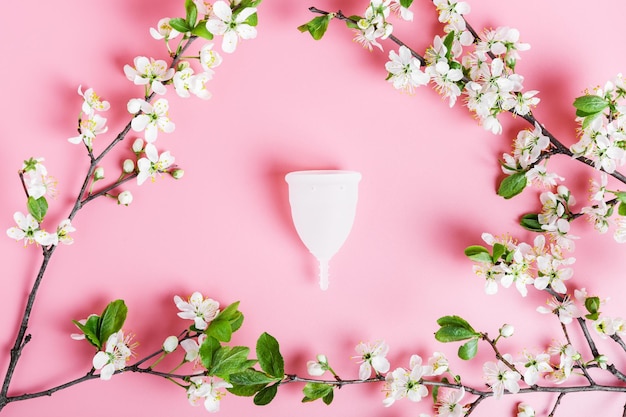 Wiederverwendbare weiße Öko-Menstruationstasse auf rosa Hintergrund Frühlingskirschbaumzweige mit weißen Blumen Ökologie- und Recyclingkonzept Zero Waste Women39s Hygiene Menstruation kritische Tage