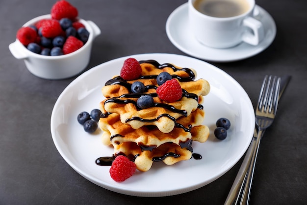 Wiener oder belgische Waffeln mit frischen Beeren Himbeeren und Heidelbeeren auf einem weißen Teller und einer Tasse Kaffee Traditionelles Dessert Closeup grauer Hintergrund