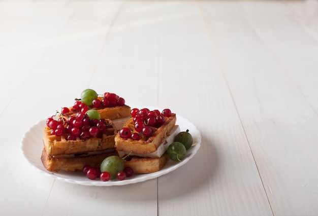 Wiener Waffeln mit roten Johannisbeeren und Stachelbeeren liegen auf einer Platte auf einem weißen Holztisch