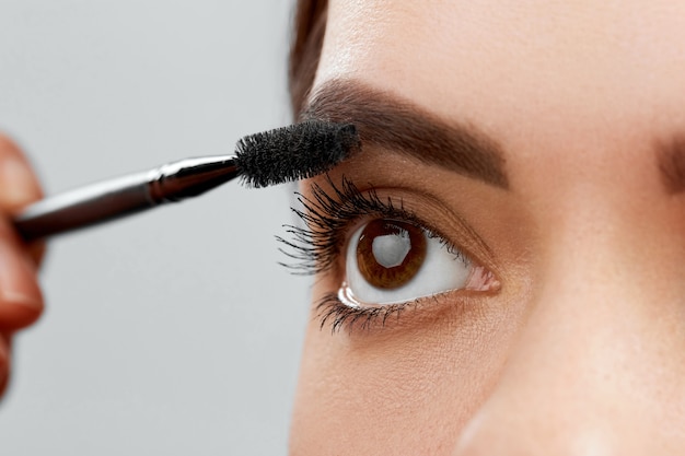 Wimperntusche. Nahaufnahme des schönen jungen Frauengesichtes mit Schönheits-Make-up, frischer weicher Haut und langen schwarzen dicken Wimpern, die Wimperntusche mit kosmetischem Pinsel anwenden.