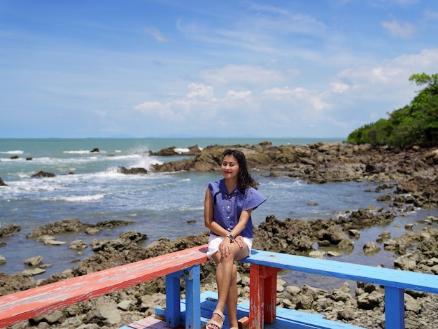 Windige Haarfrau Touristenfrau sitzt auf roter und blauer Holzsitz-Terrassenecke mit Felsenstrand-Meerblick und strahlend blauem Himmel in Ban Hua Laem, Chanthaburi, Thailand