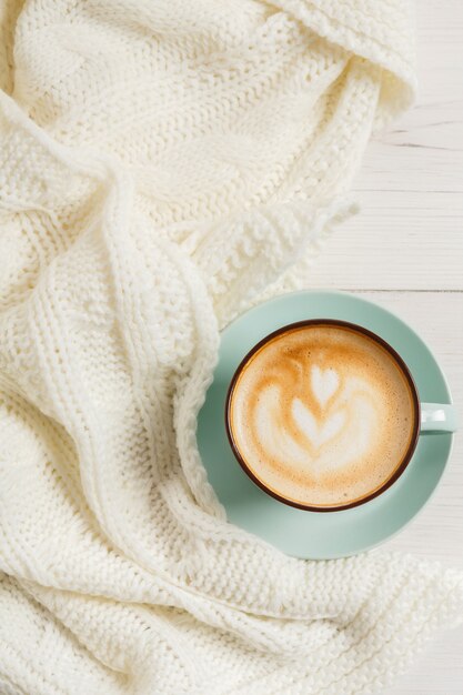 Winter Cappuccino Zusammensetzung. Blaue Kaffeetasse Draufsicht mit Schaum und gestricktem Pullover am weißen Holztisch. Warmes Heißgetränkekonzept