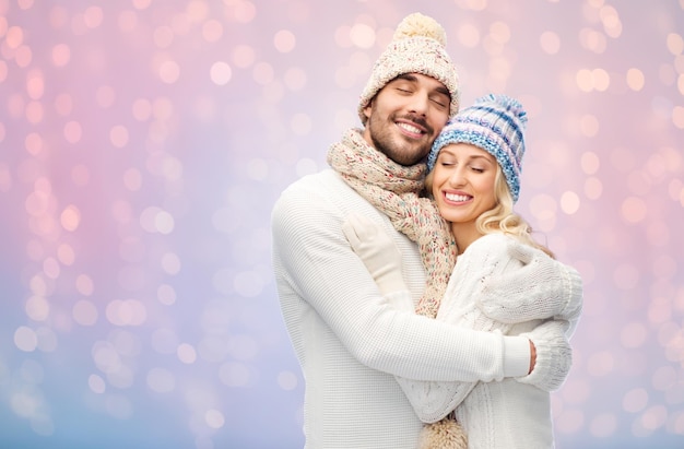 Winter-, Feiertags-, Paar-, Weihnachts- und Menschenkonzept - lächelnde Männer und Frauen mit Hüten und Schal, die sich über Rosenquarz und heiterem Lichterhintergrund umarmen