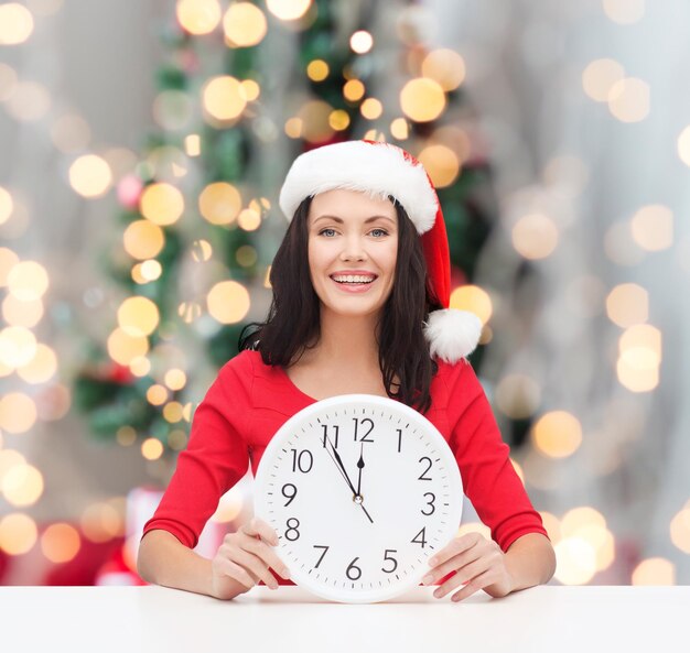 Winter-, Feiertags-, Zeit- und Leutekonzept - lächelnde Frau im Sankt-Helferhut und im roten Kleid mit Uhr über Weihnachtsbaum beleuchtet Hintergrund