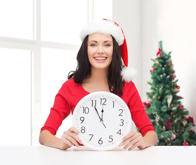 Winter-, Feiertags-, Zeit- und Personenkonzept - lächelnde Frau in Sankt-Helfermütze und rotem Kleid mit Uhr über Wohnzimmer mit Weihnachtsbaumhintergrund
