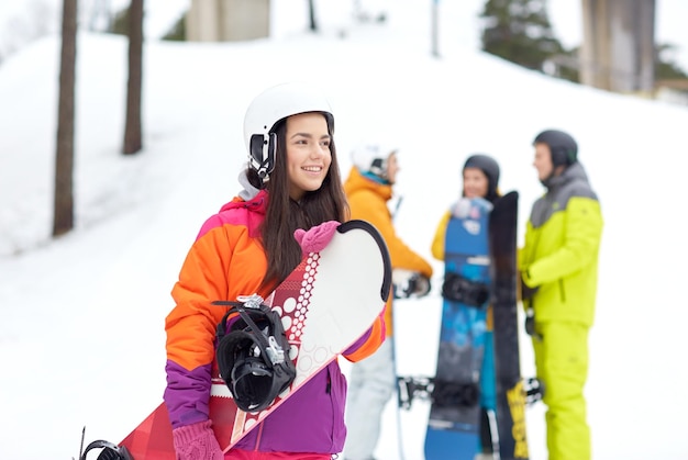 Winter-, Freizeit-, Extremsport-, Freundschafts- und Menschenkonzept - glückliche junge Frau im Helm mit Snowboard und Gruppe von Freunden